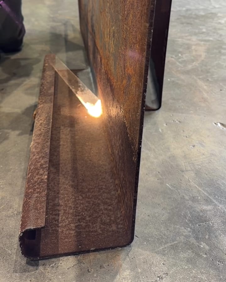 A laser cutting a bright stripe of bare metal through heavy rust on a steel angle, the ablation spot glowing where the beam meets the surface